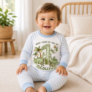 Baby wearing personalised dinosaur first birthday pyjamas with custom name and age sitting on bed